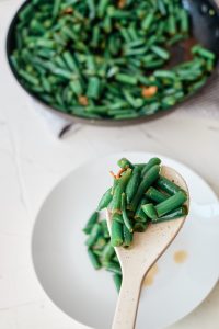 A wooden spoon holding a serving of green beans up close, over a white plate. Sauté pan of green beans in the background