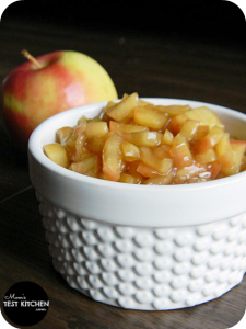 A white bowl filled with diced apples coated in a cinnamon brown sugar mixture. A red apple is in the background
