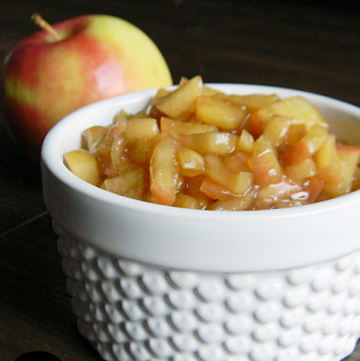 A white bowl filled with diced apples coated in a cinnamon brown sugar mixture. A red apple is in the background