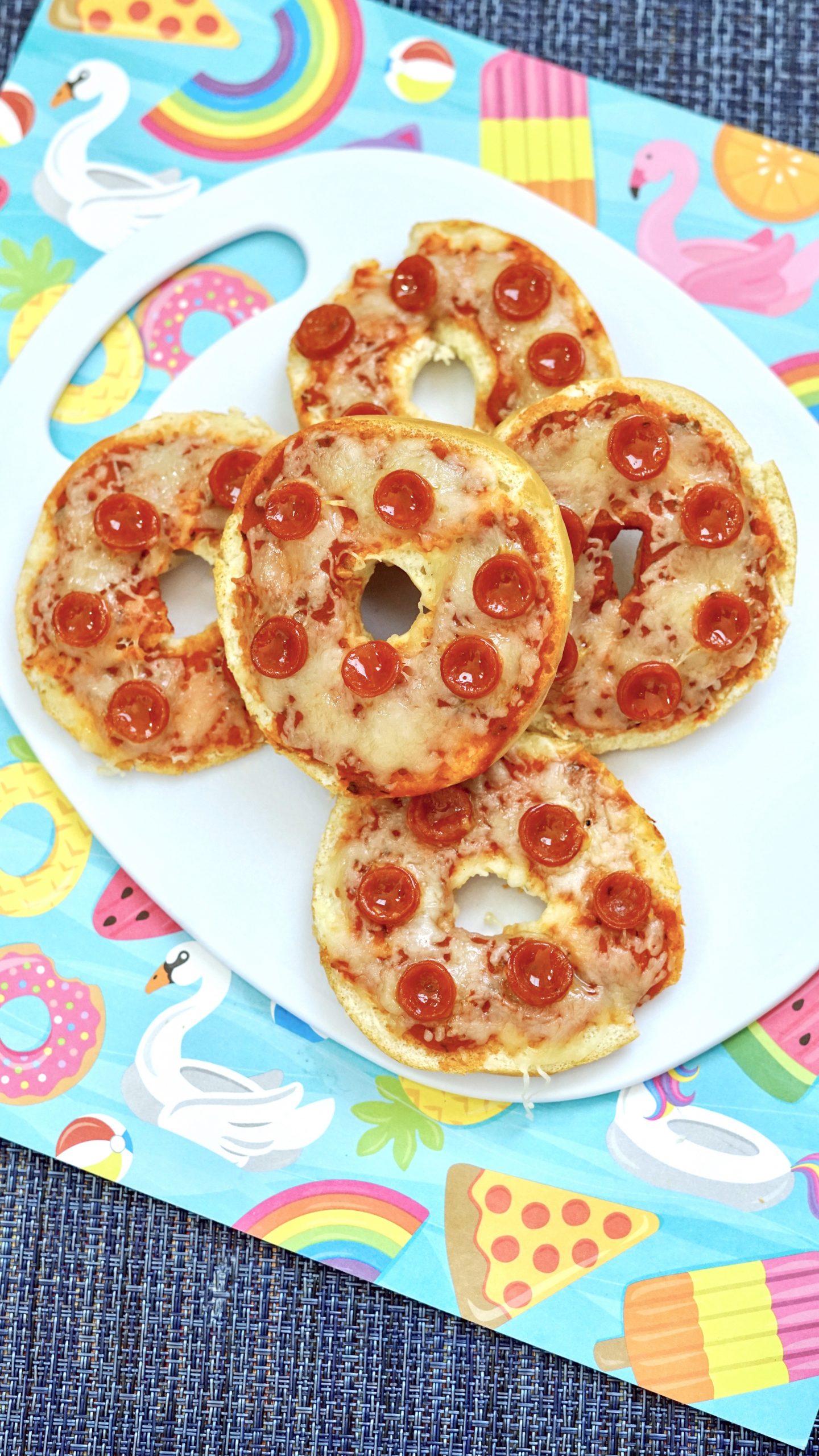 Five Air Fryer Pepperoni Bagel Bites stacked, sitting on top of a white plate that is on top of a kid-friendly, multi-colored placemat.