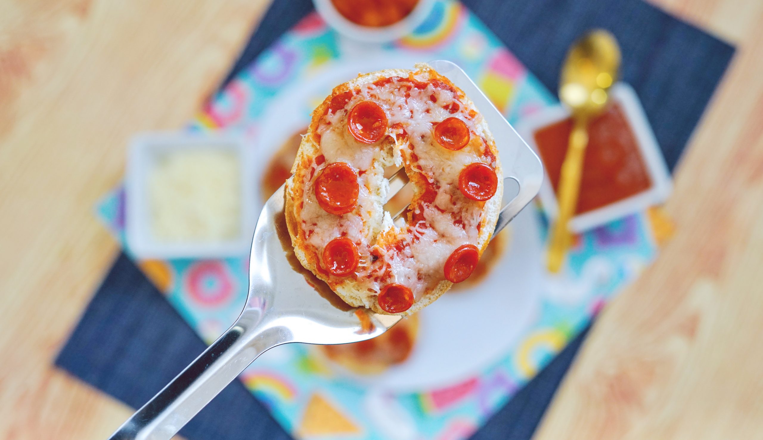 an air fryer pepperoni bagel bites on a spatula in the foreground. A dark blue placemat, and a multi-colored kid-friendly placemat in the background, with bowls of cheese and pizza sauce