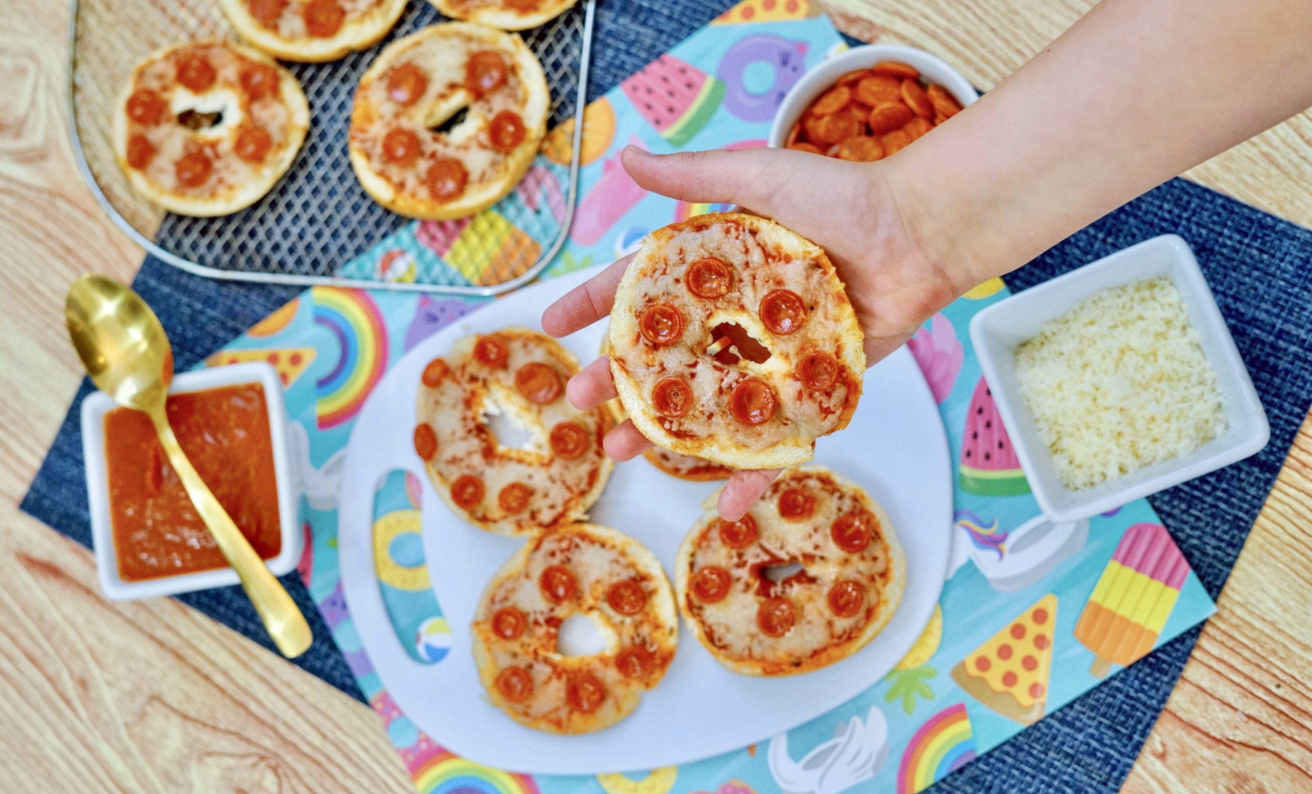 A childs hand holding an pepperoni bagel bite in the foreground. 3 Bagel bites on a white plate in the background, with tomato sauce in a bowl with a spoon. Mozzarella cheese in a bowl. An Air fryer rack with three more bagel bites sitting next to the plate.