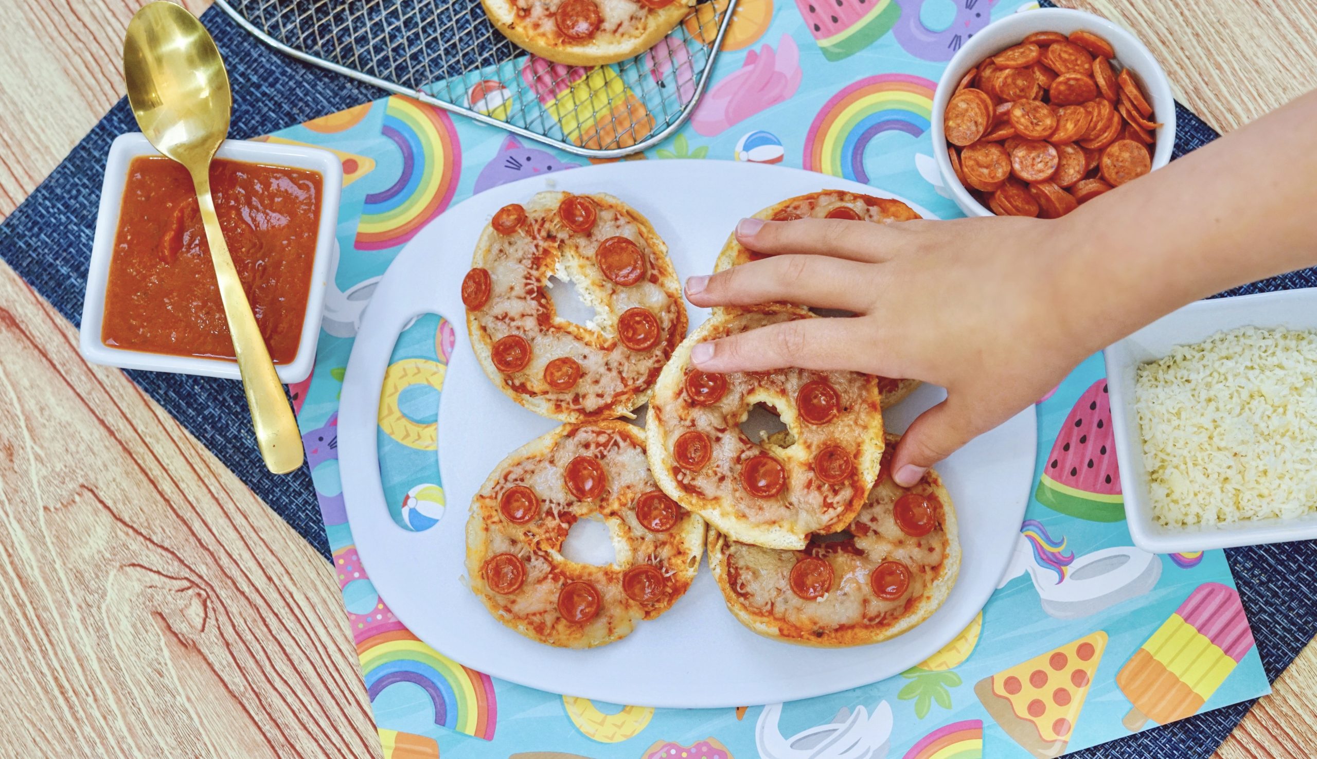 A childs hand grabbing a pepperoni bagel bite in the foreground. 3 Bagel bites on a white plate in the background, with tomato sauce in a bowl with a spoon. Mozzarella cheese in a bowl. An Air fryer rack with three more bagel bites sitting next to the plate.