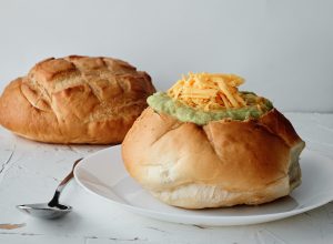 A bread bowl sitting on a white plate, filled with soup and topped with cheddar cheese. A silver dinner spoon is next to the plate. Another loaf of bread for a bread bowl is sitting in the background
