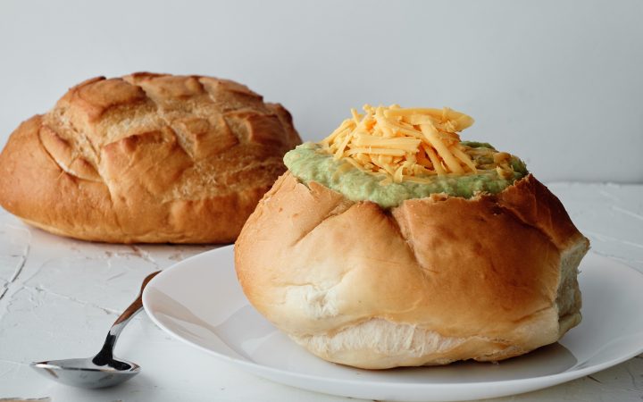 A bread bowl sitting on a white plate, filled with soup and topped with cheddar cheese. A silver dinner spoon is next to the plate. Another loaf of bread for a bread bowl is sitting in the background