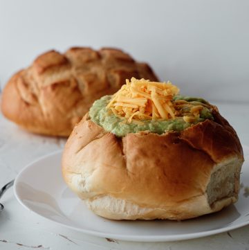 A bread bowl sitting on a white plate filled with Broccoli Soup topped with shredded cheddar cheese. A second loaf of bread in the background.