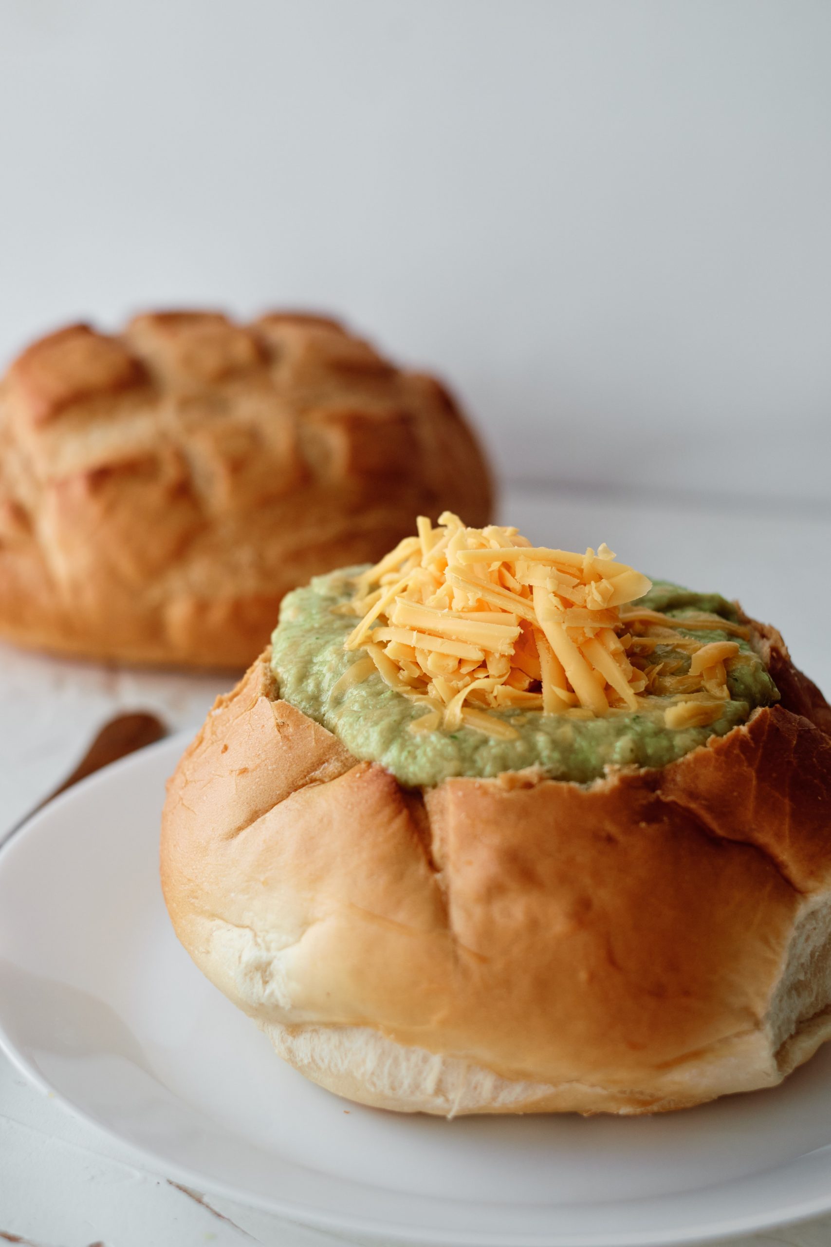 A bread bowl sitting on a white plate filled with soup topped with shredded cheddar cheese. A second loaf of bread is sitting in the background.