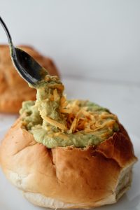 A bread bowl filled with soup, topped with shredded cheddar cheese, with a silver spoon picking up a scoop