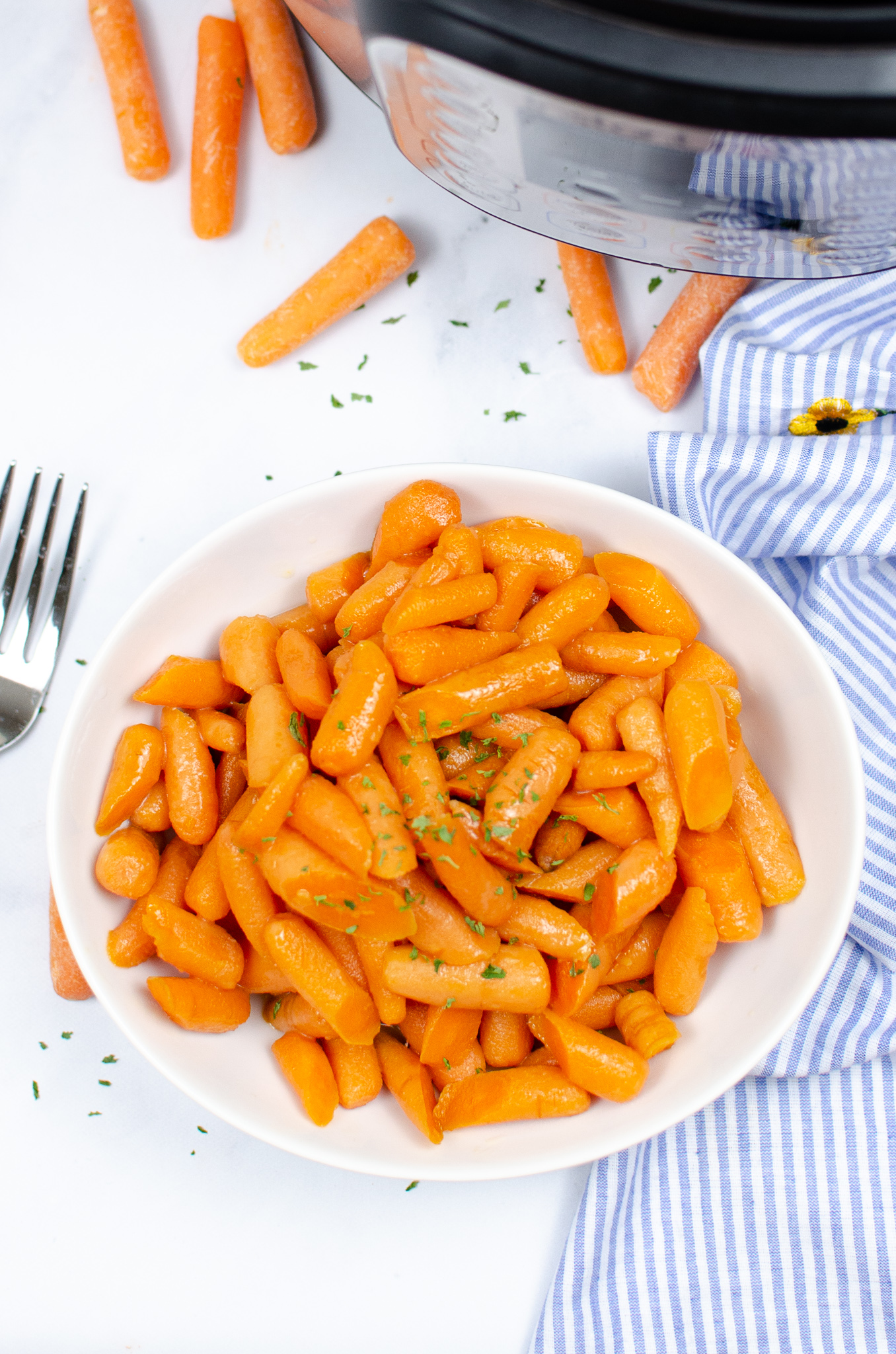 white plate with baby carrots topped with parsley sitting next to a fork and white and blue striped towel. Instant Pot in background