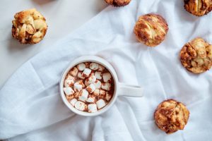 a cup of hot chocolate with marshmallows sitting on a white cloth surrounded by muffins