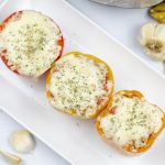 3 stuffed peppers sitting on a white rectangular plate with garlic cloves and a mustard and white polka dot towel