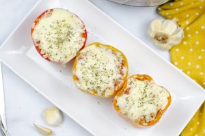 3 stuffed peppers sitting on a white rectangular plate with garlic cloves and a mustard and white polka dot towel