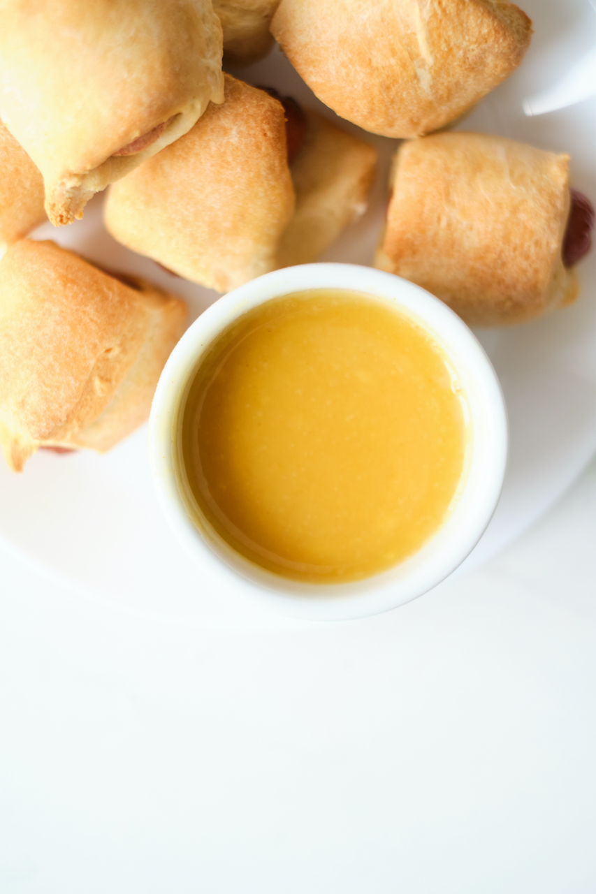 a small white bowl filled with homemade honey mustard dressing sitting on a white plate with pigs in a blanket