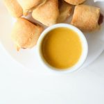 a white bowl of homemade honey mustard sitting on a white plate with pigs in a blanket