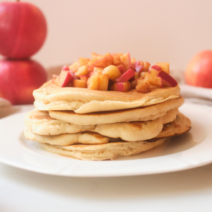 a stack of apple cinnamon pancakes sitting on a white plate with fried cinnamon apples sitting on top. Whole red apples in the background