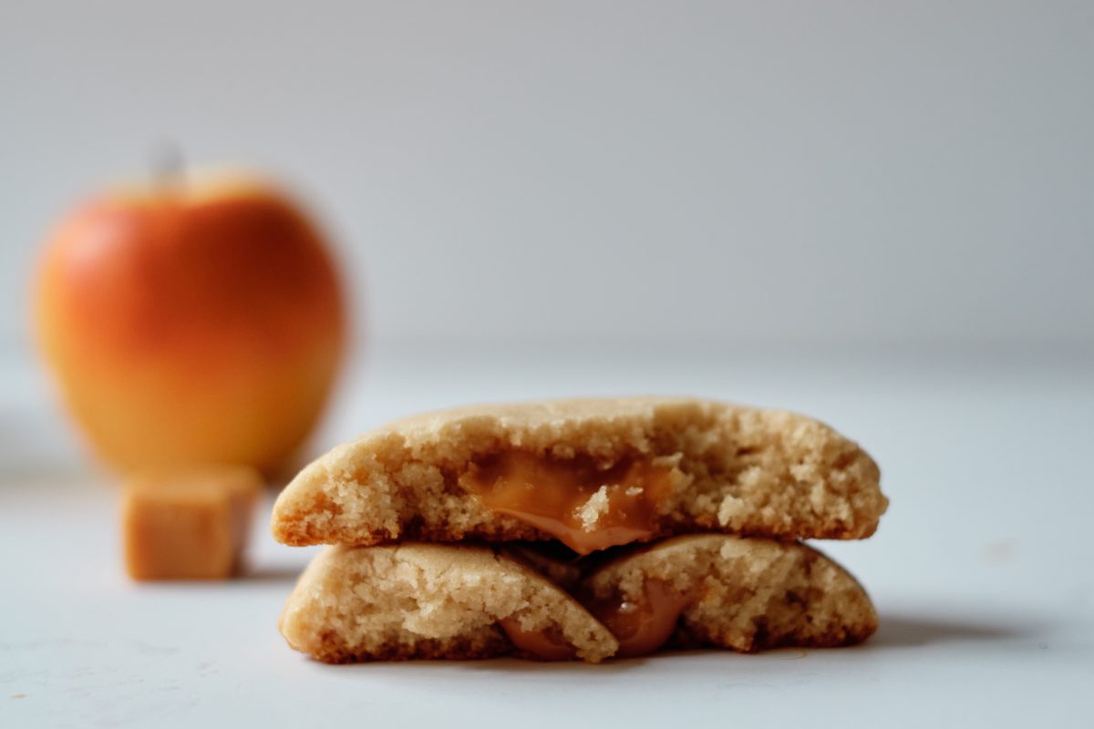 one cookie torn in half, stacked on top of each other, showing the warm, melted caramel in the middle of the cookie. a whole apple and caramel blurry in the background