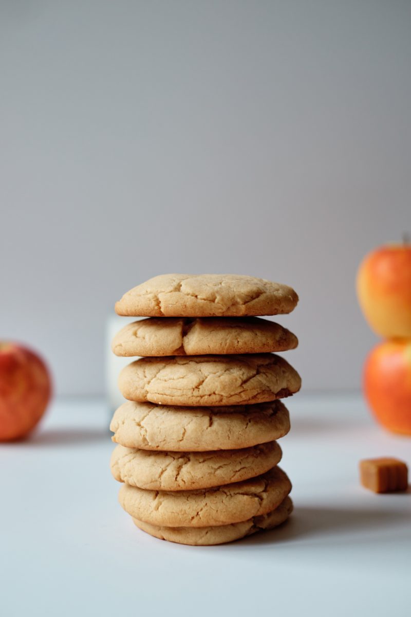 a stack of 7 cookies with whole red apples, caramel squares, and a glass of milk in the background